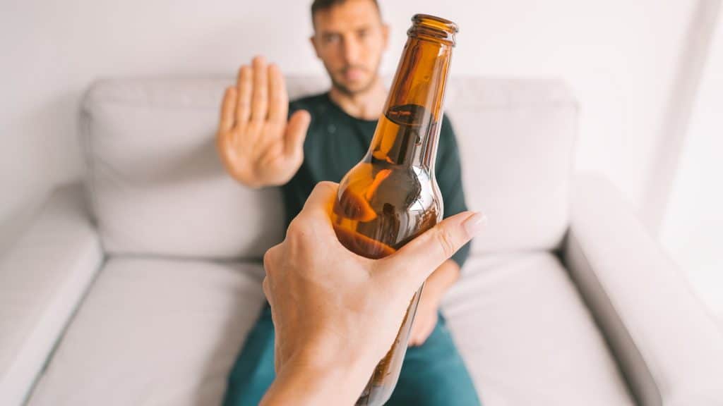 Man sitting on a couch rejecting a beer bottle with a raised hand, symbolizing alcohol detox and recovery efforts in Arizona adults.