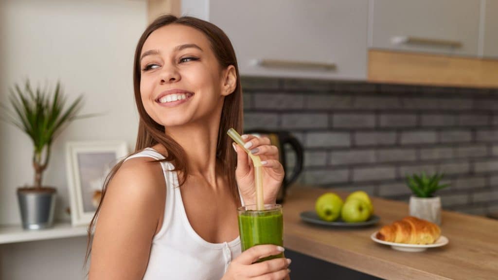 Smiling woman drinking a green detox smoothie in a kitchen, symbolizing a healthy lifestyle and recovery during alcohol detox.