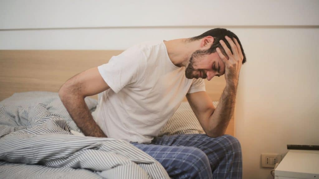 Man sitting on bed holding his head in pain, showing symptoms of an alcohol hangover and need for recovery tips.