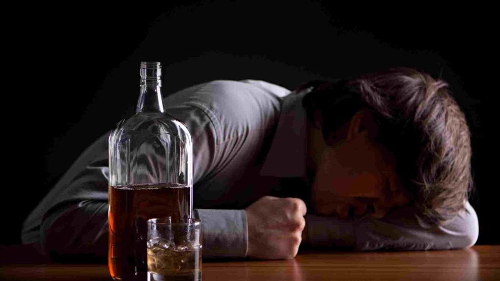 Man slumped over table, clutching throat, with a bottle of alcohol and glass beside him.