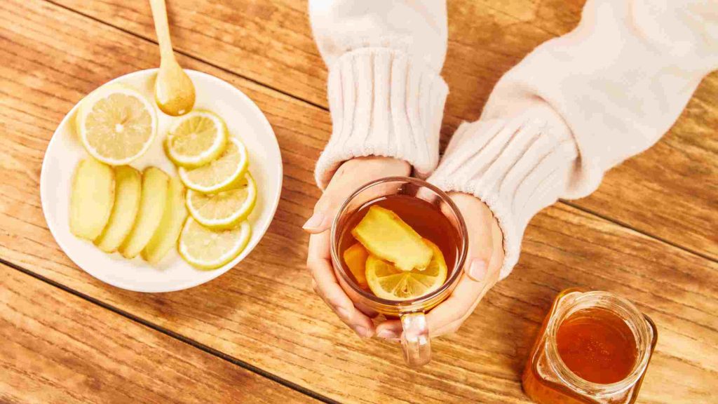 Hands holding mug of honey and lemon tea next to sliced lemons, ginger, and a jar of honey.