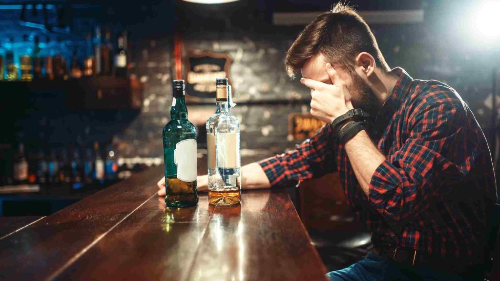 Bearded man at bar resting forehead in hand beside two nearly empty bottles.