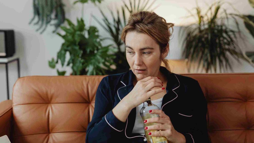 Middle-aged woman on couch holding a bottle, looking unwell and contemplative.