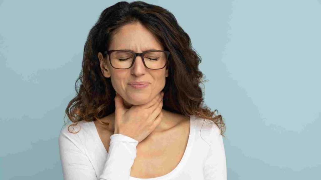 Woman closing eyes and holding her throat in discomfort against a plain background.