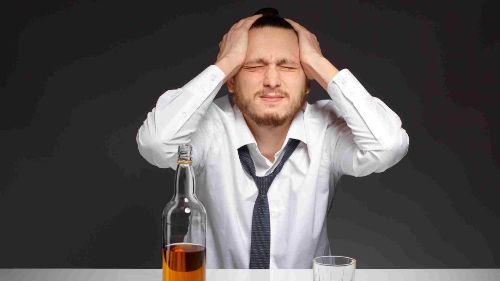 Tense man clutching his head over a bottle and glass, eyes closed in discomfort.