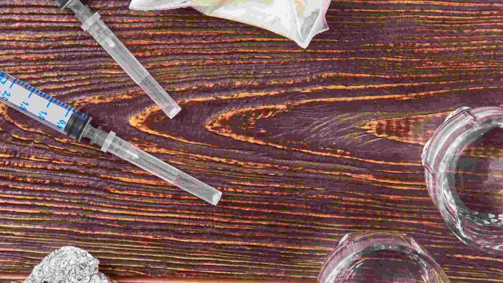 Syringes, foil, and glass items on wooden surface, showing heroin being wrapped in foil, heated, and inhaled as vapour.
