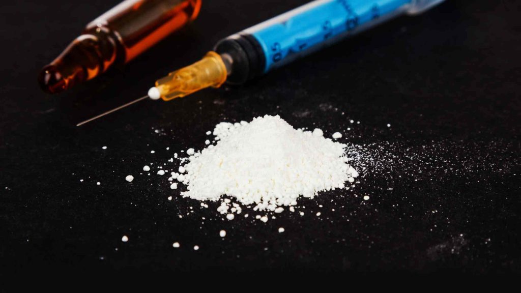 Pile of white powder heroin with syringe and ampoule on black surface, representing an overview of the drug and its risks.